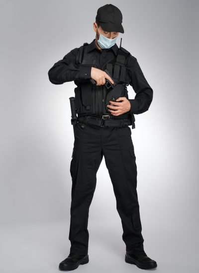 Confident policeman in uniform hiding handgun in holster indoors. Serious police officer in protective mask looking down, while removing gun, on gray studio background. Concept of work, quarantine.
